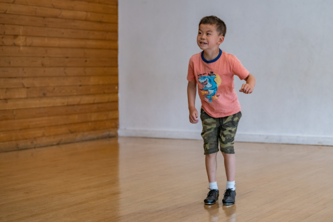 A young, smiley boy is bending his knees wearing tap shoes in a dance studio.