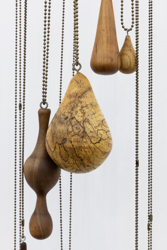Wooden teardrop-shaped pendants hang on metal chains from a large ring, which has been mounted on a white wall.
