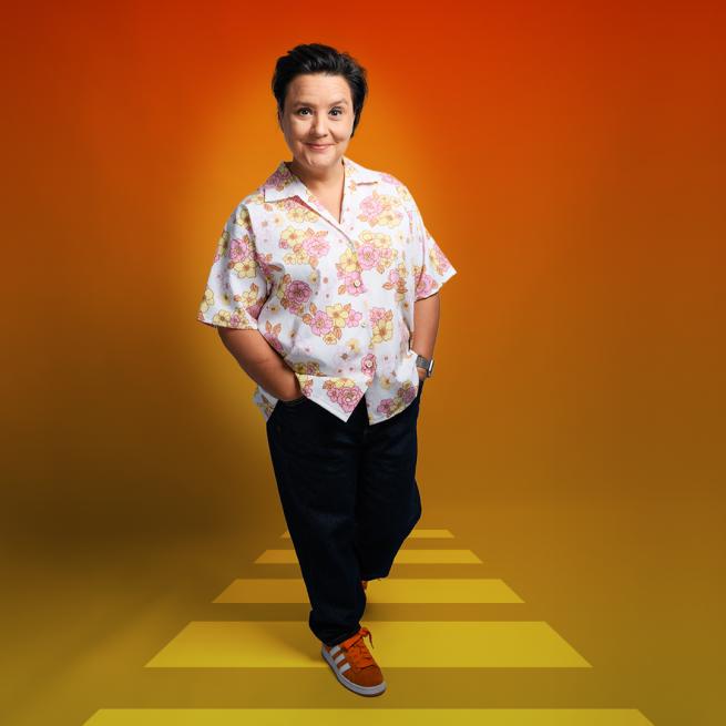 Susan Calman casually stands smiling, with hands in pockets, wearing a floral white shirt and red trainers against an orange backdrop.