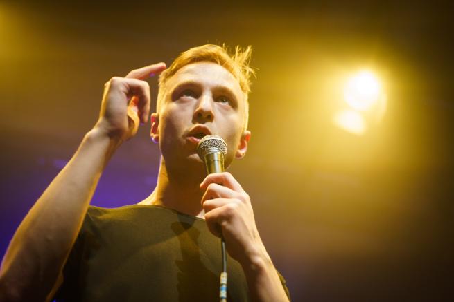 A low-angle shot of a man speaking or performing into a microphone on a stage. He has a focused expression and is gesturing with one hand, illuminated by a warm, bright yellow spotlight