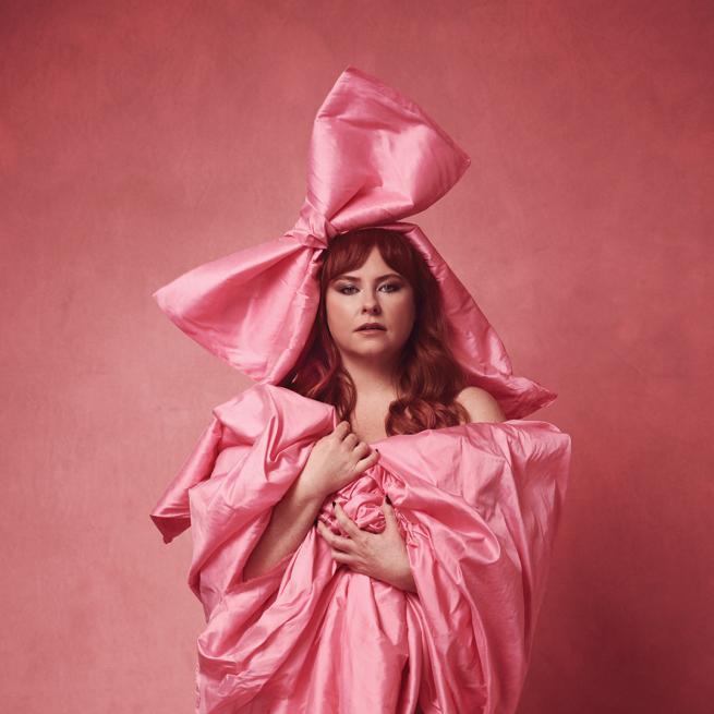 Amy Geldhill wrapped in an oversized pink bow and layers of pink, silky fabric. She holds a neutral expression and looks at the camera.