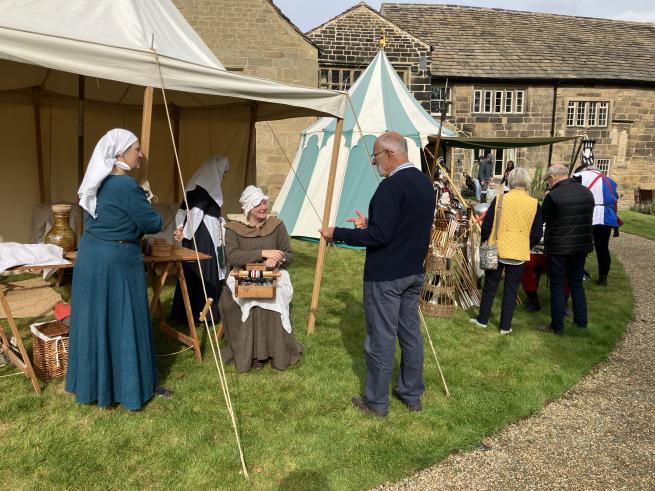 Historical Re-enactors dressed as Tudor era people sit in medieval style tents speaking to visitors. The tents are on a lawn in front of Tudor medieval manor house, Calverley Old Hall.