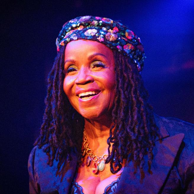 P. P. Arnold smiles candidly on stage, wearing a a sequined beret-style cap and textured suit jacket. She is lit by a warm stage light and there is a hazy blue hue in the background.