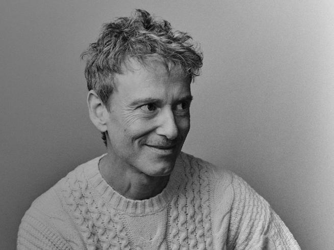 A man with short light hair, wearing a white jumper smiling.