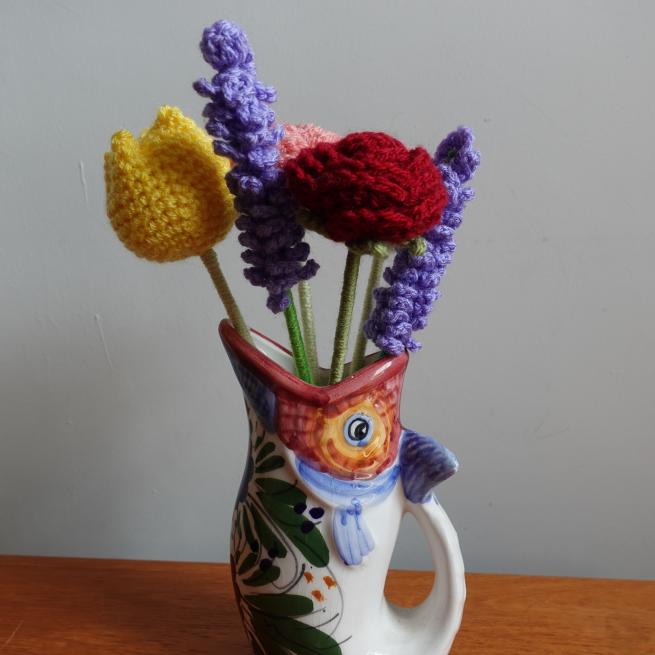 A photograph of 5 crochet flower stems in a vase shaped like a fish. The flowers are yellow, purple, red and pink.