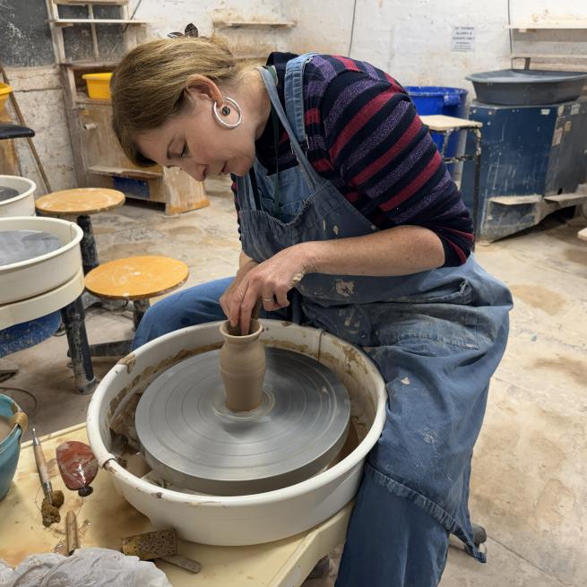 A woman is sat at a pottery wheel and is throwing a small vase
