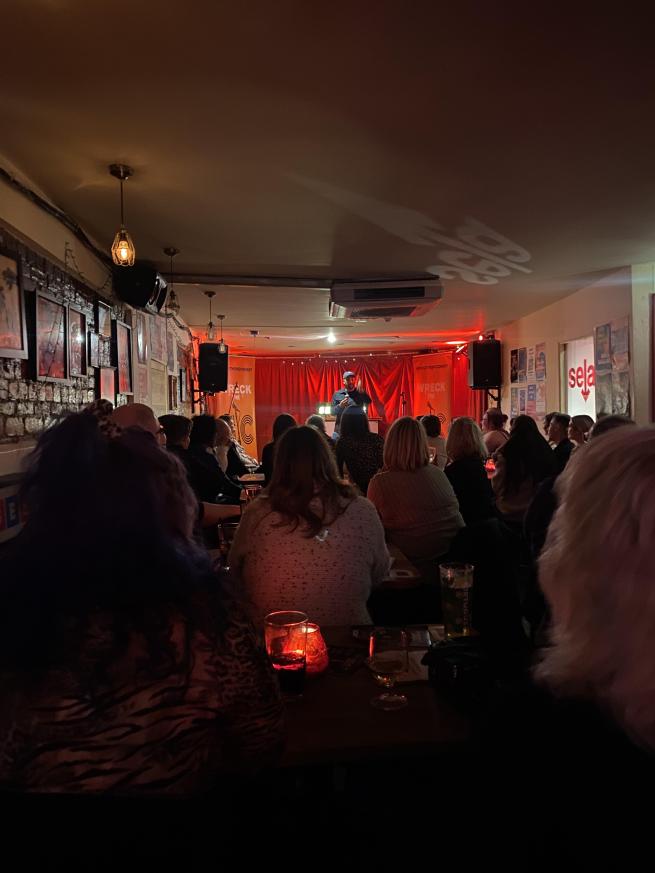 A man with a microphone stands on stage to a packed out bar with lots of people watching.
