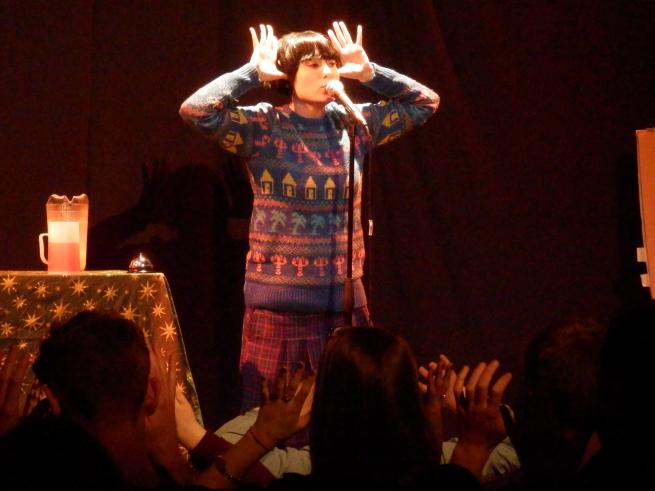 A person stands on a stage with their hands splayed at either side of their head, an audience is in the foreground making the same gesture.