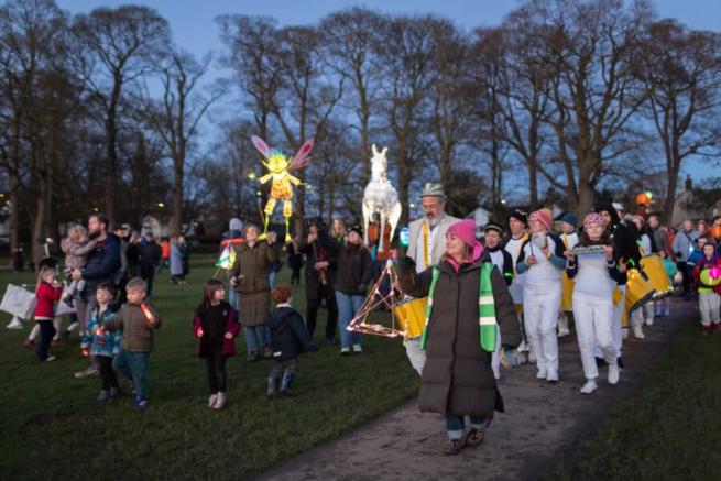 Group of people are walking down a path in park holding lanterns which are lit up. There is a sprite and horse lantern and a Bataria playing drums.