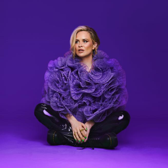 Joanne McNally looks to the side and sits cross-legged in shiny black trousers and an oversized, frilly purple top, in front of a purple background.