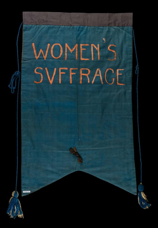 A hand made banner reads "Women's Suffrage" in orange writing on green fabric