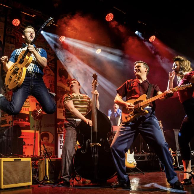 Live production shot of musicians performing on stage, with a guitarist leaping in the air beneath white and red spotlights.