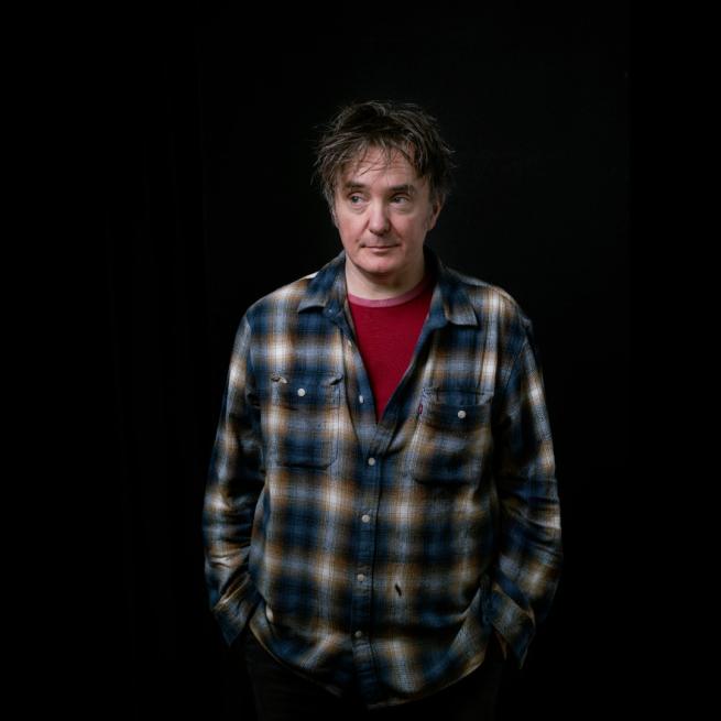 Dylan Moran stands against a black backdrop, relaxed with hands in his pockets and wearing a plaid shirt and red undershirt.
