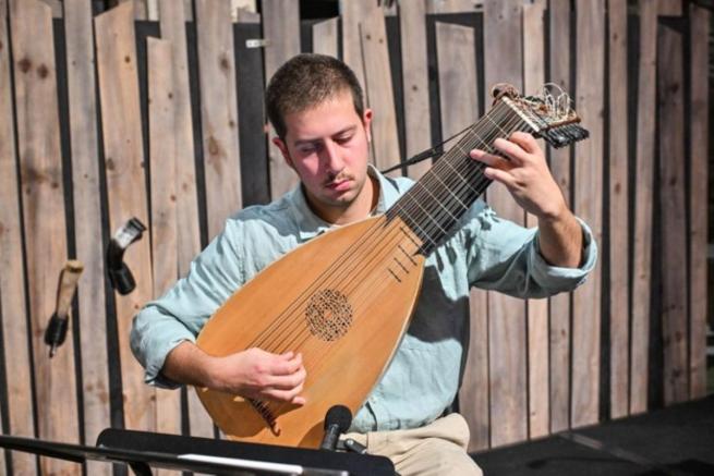 A man in a blue shirt sits in front of a fence, he is playing a lute.