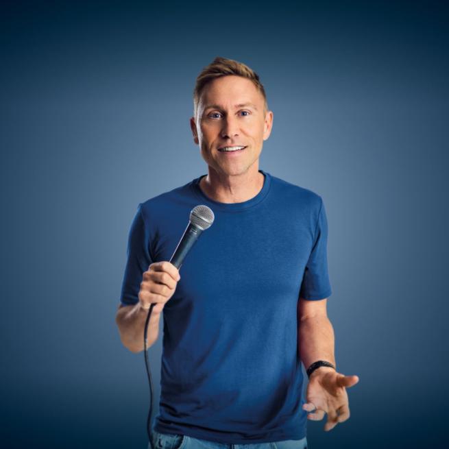 Russell Howard in a blue t-shirt holds up a microphone and smiles at the camera.