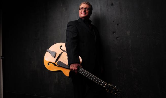 Man in Black jacket and trousers standing in front of a black background, he's holding an acoustic guitar while staring into the distance in a thoughtful way