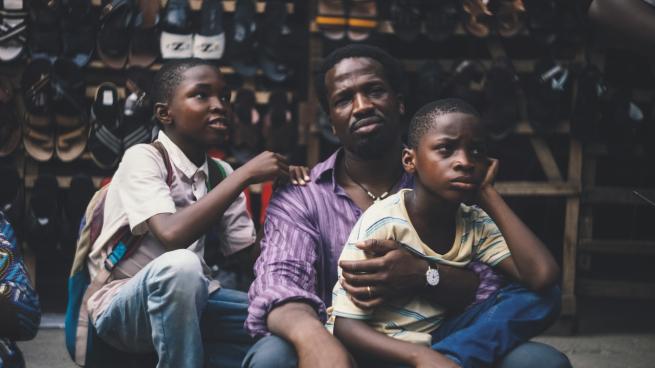 A man sits with two children resting against him while another child sits nearby. They are seated outdoors in front of a display of shoes arranged on shelves.