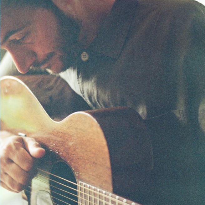 Grainy close-up shot of Tom Smith playing an acoustic guitar, with a subtle green hue. 