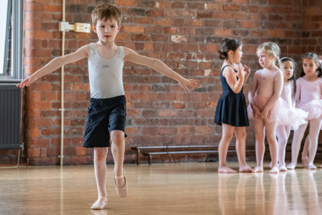 A young boy is ballancing on one leg, arms stretched, whilst a group of young girls are gathered behind him in a dance studio.