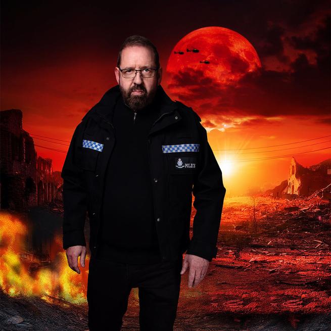 A stern Alfie Moore in police uniform stands in front of a red-tinted apocalyptic landscape, featuring flaming rubble, ruined buildings and helicopter silhouettes in the sky.
