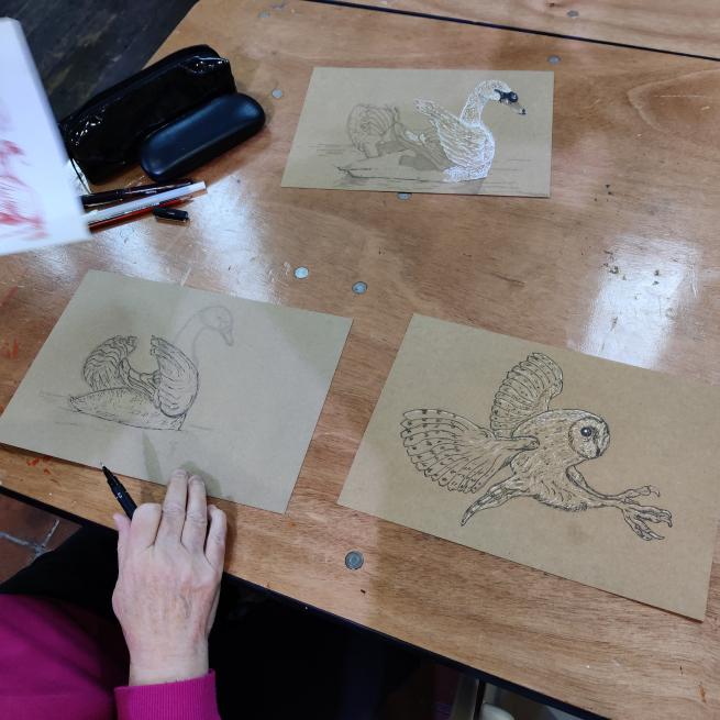 A photograph of three bird drawings lay on a wooden table. The drawing are of an owl and two swans, they are on brown paper and use white and black pens and pencils