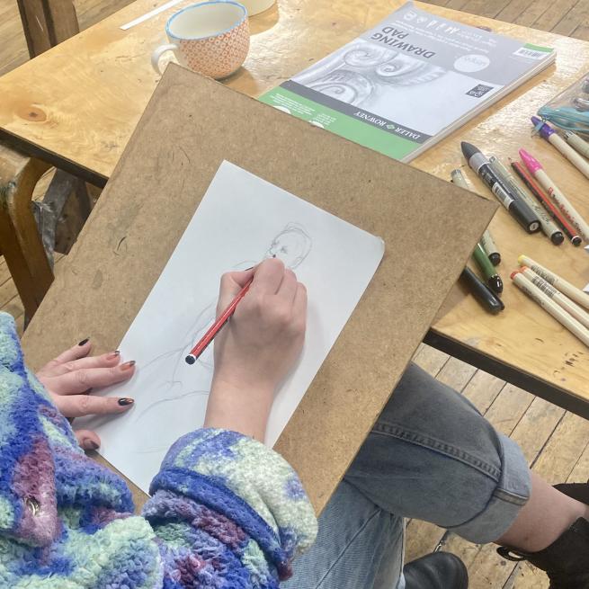 A photograph taken over the shoulder of someone with a drawing board on their knee working on a pencil drawing of a person