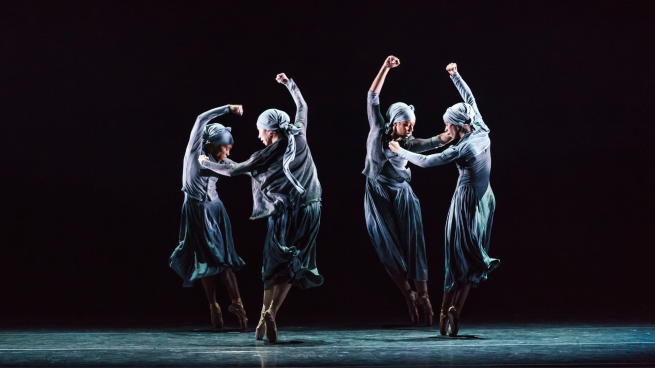 Four performers dance on stage in pairs. They have their arms in the air. 
