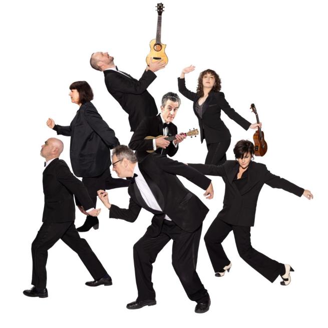 Seven people in matching black suits strut around a plain white backdrop with exaggerated movements, some holding a ukulele .