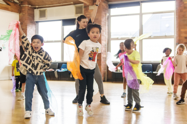 Children are dancing and jumping with different coloured scarves in a dance studio.