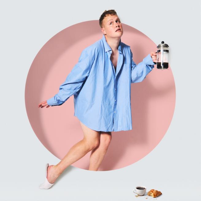 Emerging from within a pale pink circle, Josh Jones tip toes in slippers and an oversized blue shirt, holding a cafetiere. A mug of coffee and half-eaten croissant are visible in the foreground.