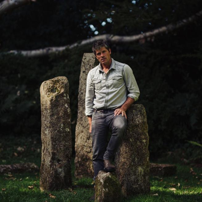 Seth Lakeman leans against a mossy standing stone in a woodland setting.