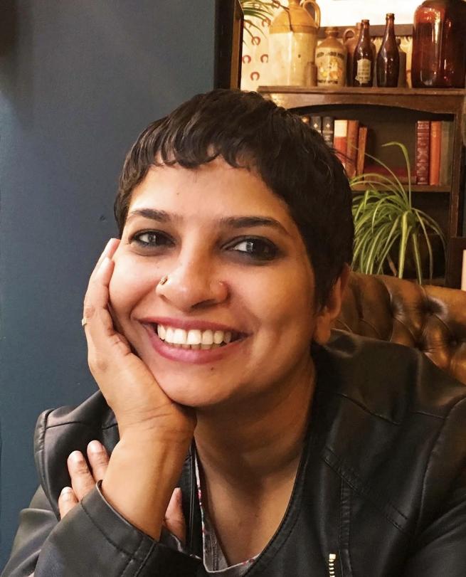 A smiling woman with short dark hair and a nose ring, resting her chin on their hand. They are wearing a black leather jacket in a room with a bookshelf and vintage bottles in the background.