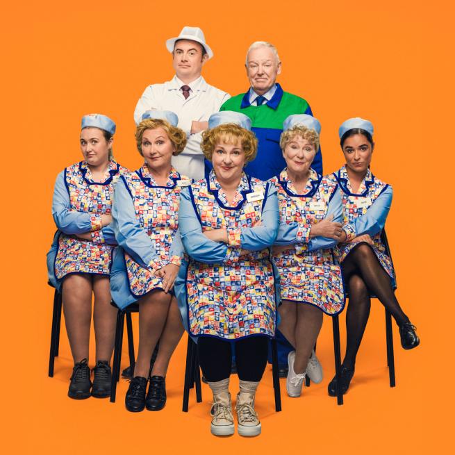 Five dinner ladies in apron uniforms sat in a row with folded arms. Behind them stand a man in blue overalls and a man in a white apron and hat.