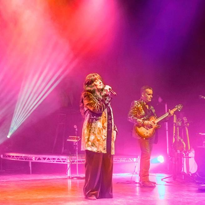 On stage, there is female singer and guitarist. Stage lights are hazy and pink.