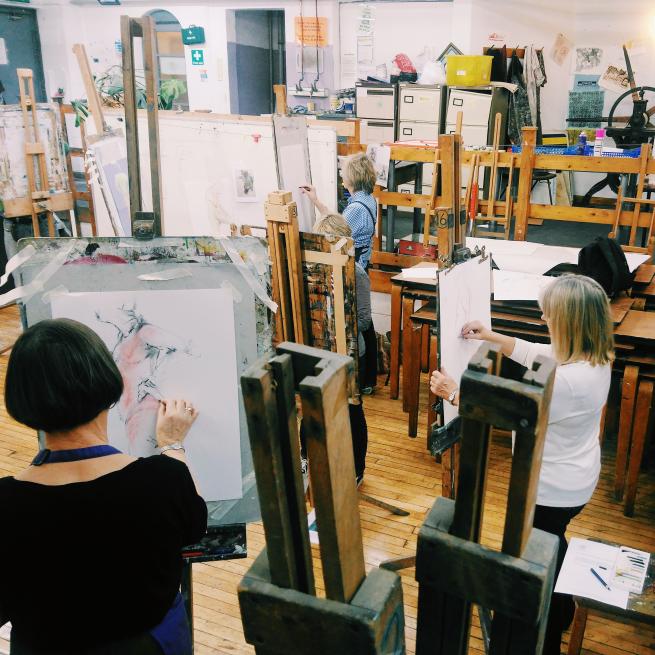 A photo of 3 women stood up at easels, they are all drawing a life model. the woman in the foreground is using a pink pencil to add colour to her work