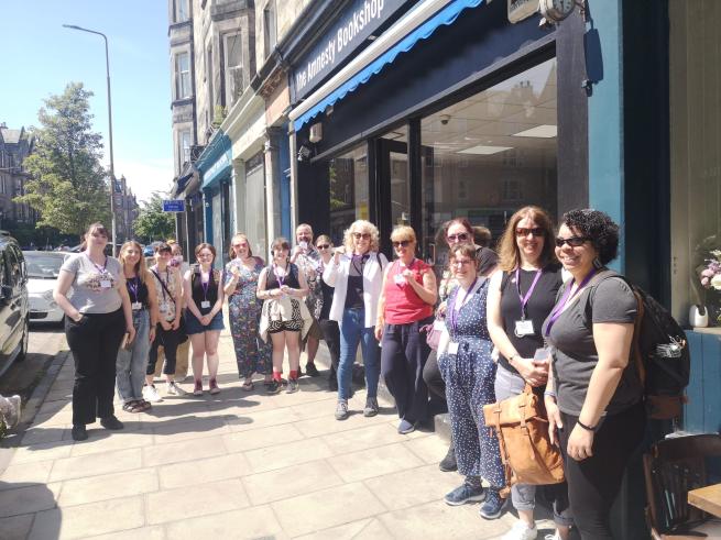 A mixed group of people standing on the pavement outside a bookshop in the sunshine.
