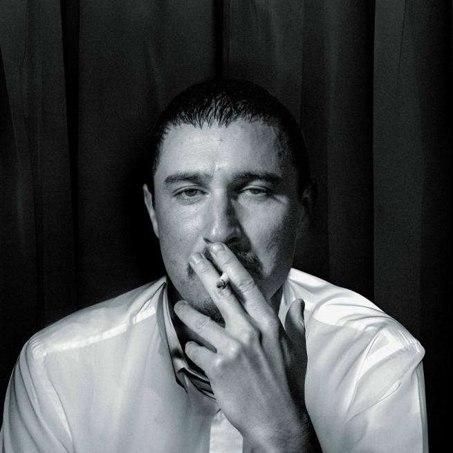 Black and white image of Tom Rosenthal smoking in the style of the Arctic Monkeys' album cover for Whatever People Say I Am, That's What I'm Not.