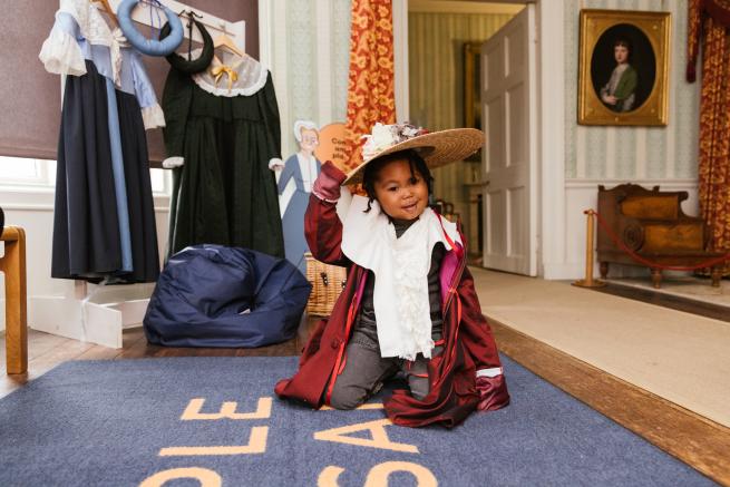 A toddler kneeling on the floor in fancy dress wearing a hat