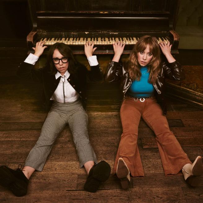 Flo and Joan sit on the floor with their legs extended, and their arms playing a piano positioned behind their heads.
