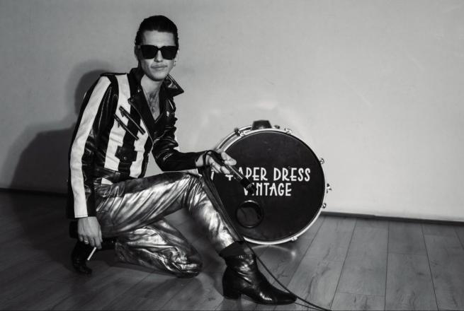 A black and white picture Tiss, wearing dark sunglasses and shiny trousers. Posing with a cigarette and drum.