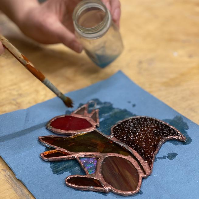 A photograph of a stained glass squirrel on some blue paper with a hand poised over it with a paintbrush  