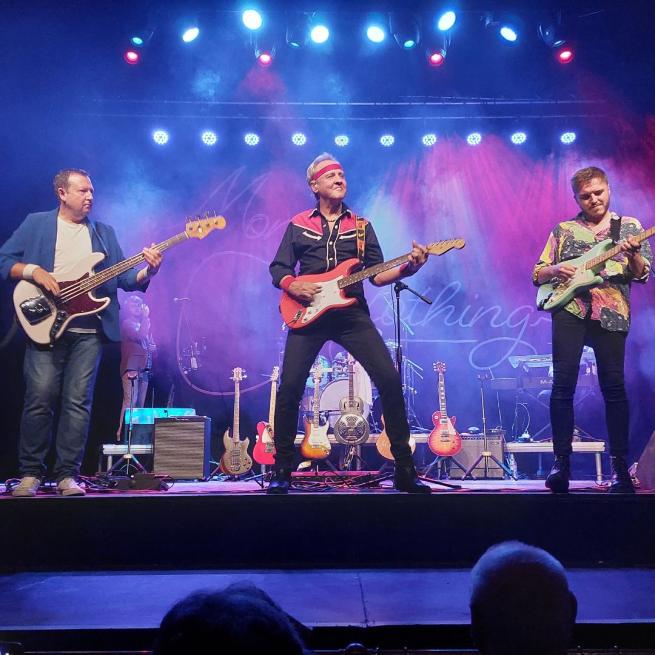Three guitars perform on stage, standing in a row, each playing electric guitars. 