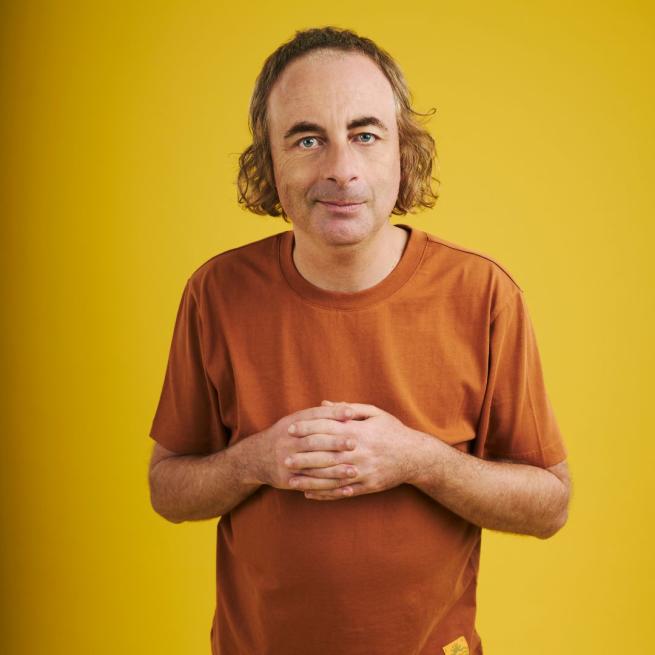 Paul Foot smirks and clasps his palms together, wearing an orange t-shirt in front of a yellow backdrop.