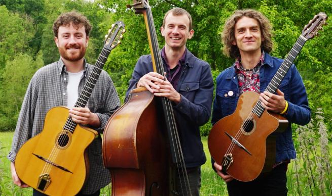 Three friendly musicians smile engagingly into the camera, two are holding guitars and the third a Double Bass, mind you I'm not sure why they're in a field.
