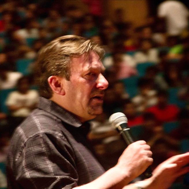 Andrew Newton performs to a packed blurry crowd, speaking into a handheld microphone and wearing a black shirt.