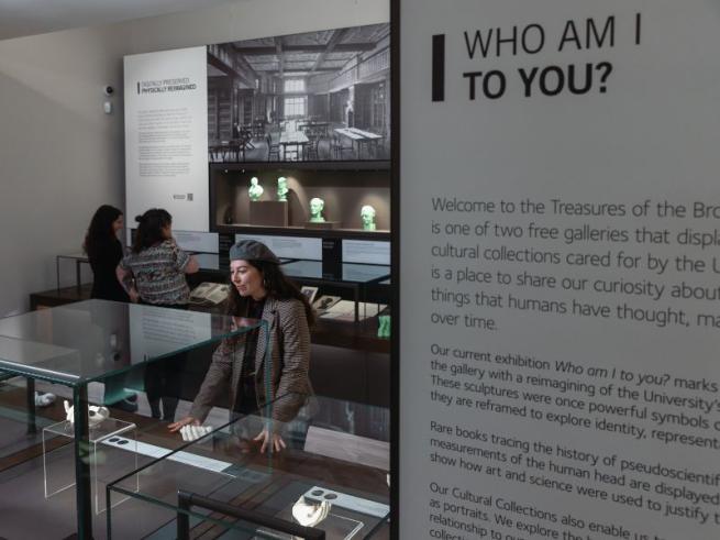 3 people stand in Treasures of the Brotherton museum. In the foreground there is an interpretation board that read 'Who am I to you?'  