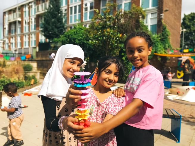 Three smiling children stand together outdoors in a community garden, holding up a colourful tower made from stacked flower-shaped pieces. Apartment buildings, greenery and other children taking part in activities can be seen in the sunny background.