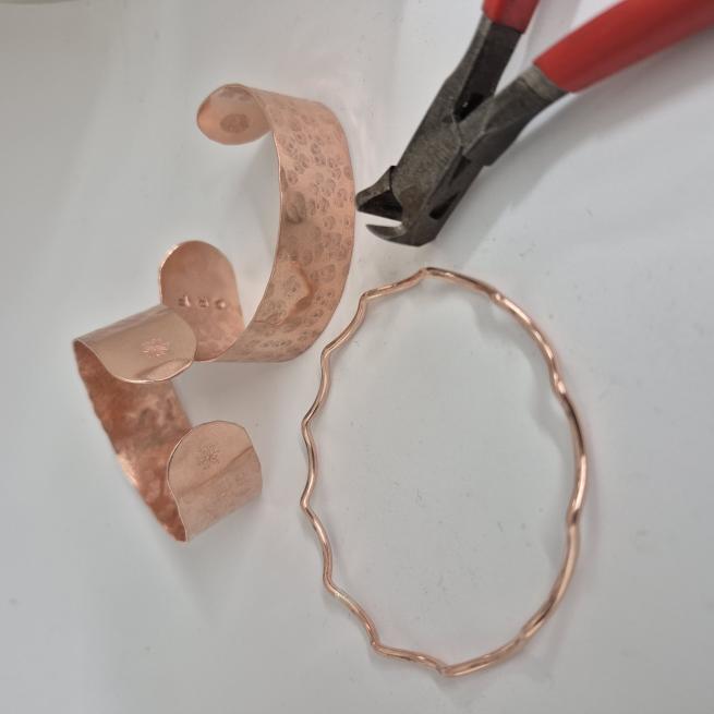 A photograph of a think copper bangle and two chunky copper cuffs lay next to a jewellery tool with a red handle