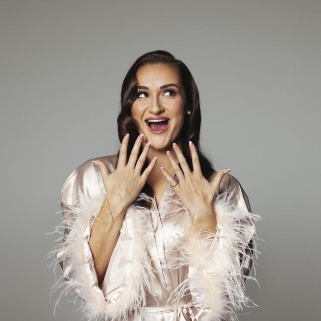 Maddie Grace Jepson smiles with an open mouth and looks off camera, holding up manicured hands. She wears a silk dressing gown with feathery sleeves and makeup.