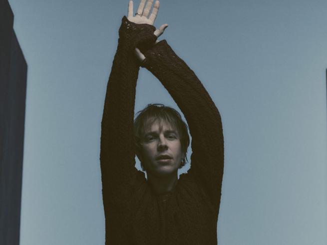 Tom Odell standing with arms raised above their head against a plain background, wearing a dark long‑sleeved top.